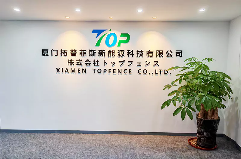株式会社トップフェンスのオフィスエントランス。壁には、ブルーとグリーンのTOPロゴと、中・日・英3言語の社名が掲げられています。隣には大きな観葉植物(パキラ)が置かれた、明るく清潔感のあるオフィスフロントです。